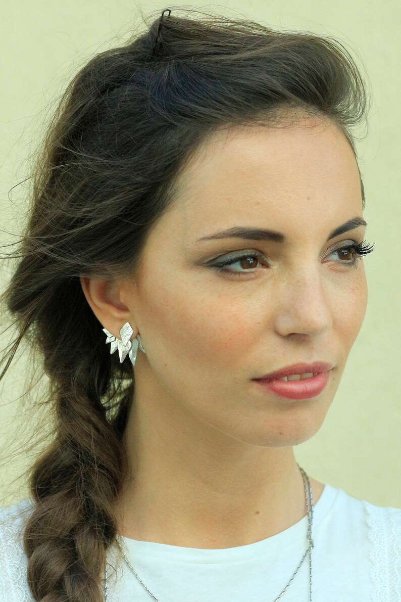 May include: A woman with long brown hair wearing a white shirt and a silver necklace. She is wearing silver earrings that are shaped like points.
