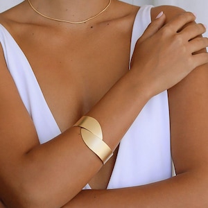 May include: A gold cuff bracelet with a curved, leaf-like design. The bracelet is worn on the wrist of a person wearing a white top.