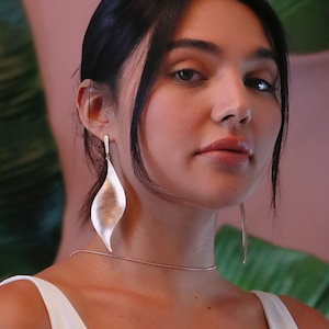 Pode incluir: Uma mulher vestindo um top branco sem mangas com um colar de corrente de ouro e brincos longos de prata em forma de folha.