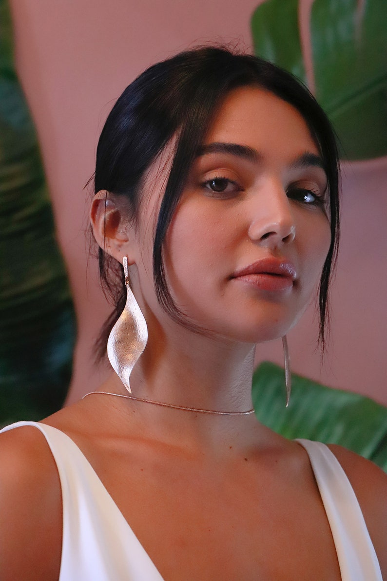 Pode incluir: Uma mulher vestindo um top branco sem mangas com um colar de corrente de ouro e brincos longos de prata em forma de folha.