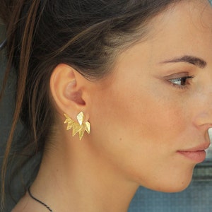 May include: A pair of gold-toned earrings with a geometric, leaf-like design. The earrings are worn by a person with long brown hair.
