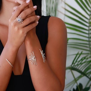 K&ouml;nnte beinhalten: Ein silberner Ring und ein Armband mit einem Sonnenstrahl-Design. Der Ring hat einen kleinen blauen Stein in der Mitte. Das Armband ist offen und hat zwei Sonnenstrahl-Designs.