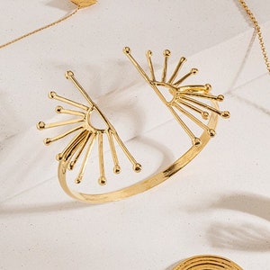 K&ouml;nnte beinhalten: Ein goldfarbenes Armband mit einem Sonnenstrahl-Design. Das Armband ist offen und hat ein breites Band. Das Sonnenstrahl-Design befindet sich auf beiden Seiten des Armbands.