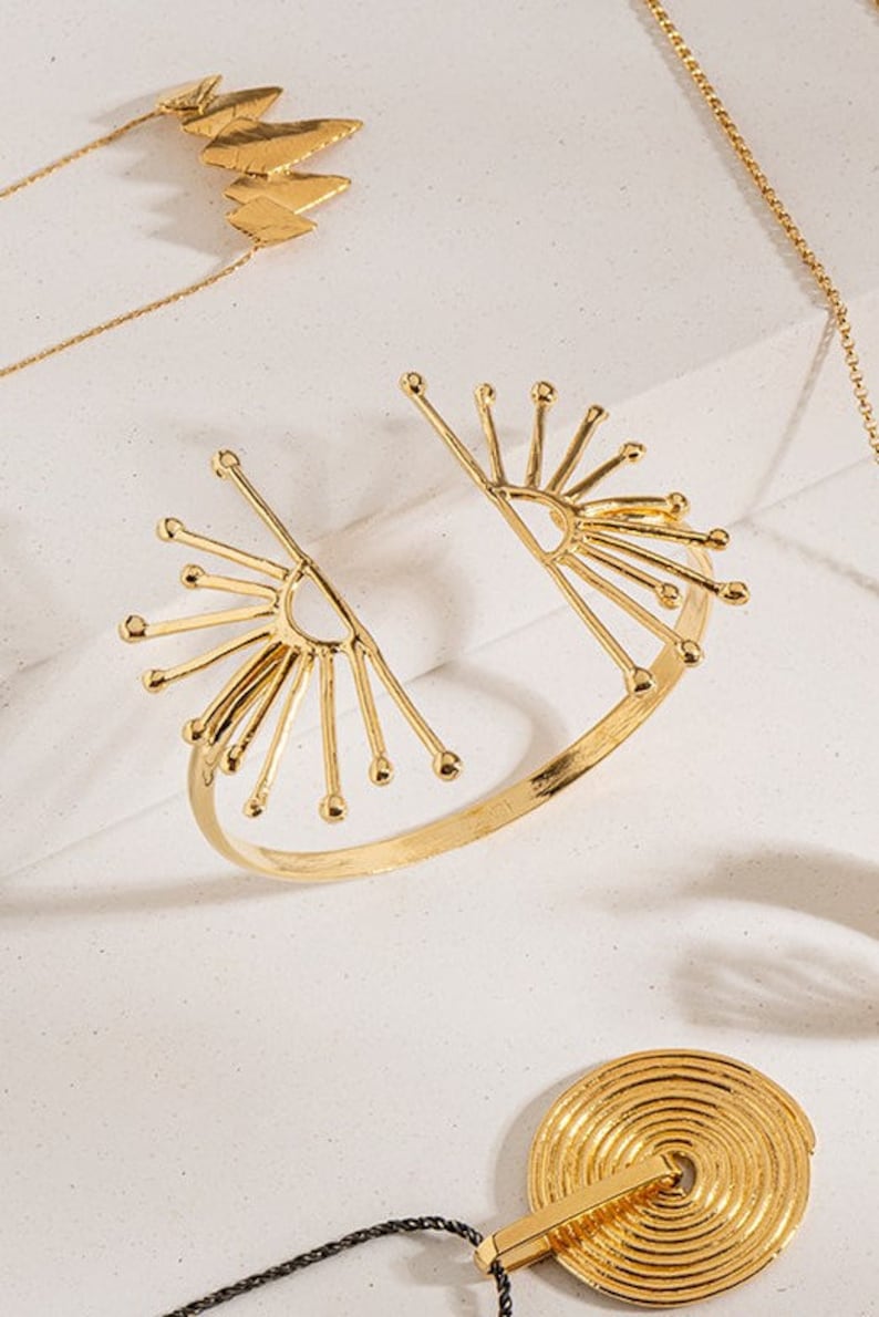 K&ouml;nnte beinhalten: Ein goldfarbenes Armband mit einem Sonnenstrahl-Design. Das Armband ist offen und hat ein breites Band. Das Sonnenstrahl-Design befindet sich auf beiden Seiten des Armbands.