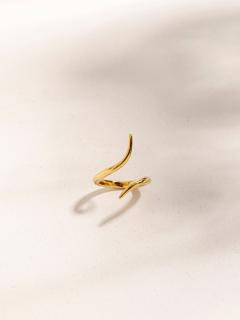 May include: A gold-colored ring with a unique, twisted design. The ring is made of metal and has a smooth, polished finish.