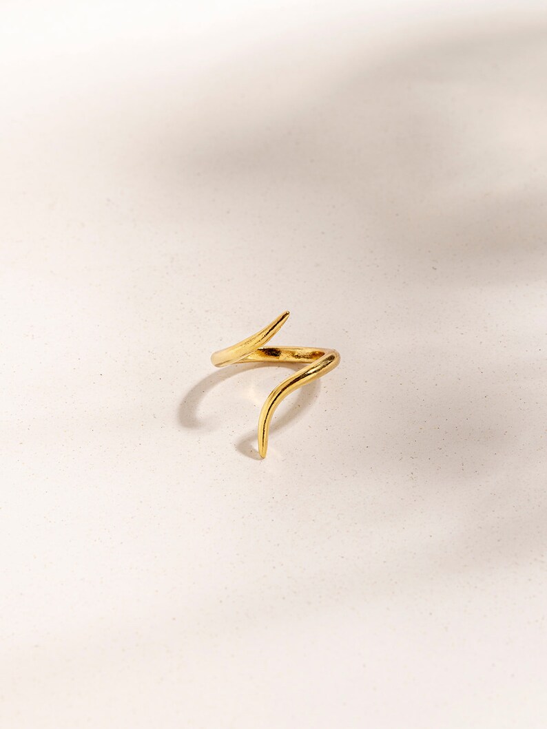 May include: A gold-toned ring with a unique, abstract design. The ring features a twisted, curved shape with a pointed end.