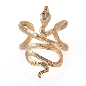 Puede incluir: Un brazalete de serpiente dorado con ojos de piedras preciosas azules. El brazalete está hecho de metal y tiene una superficie texturizada.