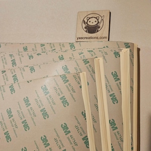 May include: A pile of adhesive sheets featuring the 3M logo and product details. A small wooden square with a cartoon character and the text "yxecreations.com" is placed on top of the stack.