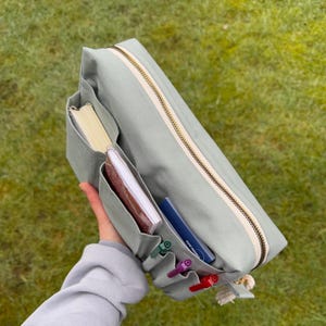 Könnte beinhalten: Hellgrünes Federmäppchen aus Canvas mit mehreren Taschen und einem goldenen Reißverschluss. Das Etui enthält Notizbücher, Stifte und Bleistifte. Vor einem grünen Gras Hintergrund.