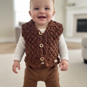 Puede incluir: Un bebé sonriente con un chaleco marrón de punto grueso con botones de madera sobre una camisa blanca de manga larga. El bebé también lleva pantalones marrones. El chaleco parece hecho a mano y añade un elemento acogedor y texturado al conjunto.