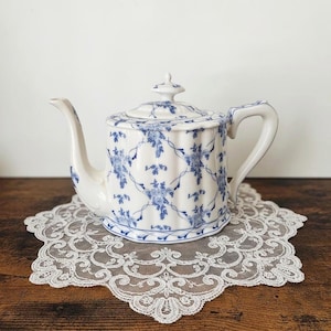 May include: A white ceramic teapot featuring a blue floral pattern with a lattice design. The teapot has a fluted body, a curved spout, a handle, and a matching lid with a small knob. The surface is decorated with intricate blue floral motifs.