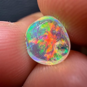 Puede incluir: Una gema de ópalo colorida con forma redondeada. El ópalo muestra un vibrante juego de colores, incluyendo tonos rojos, verdes y amarillos. Finas grietas son visibles en la superficie de la piedra.