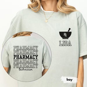 May include: A light green t-shirt with a black pharmacy mortar and pestle logo and the name "LISA". The back of the shirt features the text "PHARMACY" repeated multiple times, with "technician" below. The word "bay" is also visible.