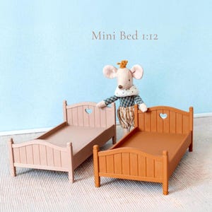 May include: Two miniature wooden beds, one pale pink and one brown, with heart-shaped cutouts in the headboards. The beds are part of a dollhouse set, with the text "Mini Bed 1:12" above them. A stuffed mouse doll is standing next to the beds.