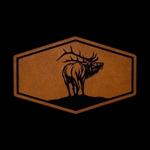 May include: A hexagonal brown leather patch featuring a black graphic illustration of an elk standing on a small hill. The patch has a dark border and a textured surface, showcasing the detailed silhouette of the animal with large antlers.