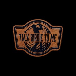 May include: A brown leather patch with the text "TALK BIRDIE TO ME" in black. The patch features a silhouette of a golfer swinging a club and a golf ball. The patch has a black border and is set against a black background.