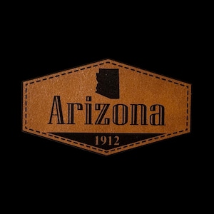May include: A tan, hexagonal leather patch featuring a black silhouette of the state of Arizona, the word Arizona in a bold serif font, and the year 1912 printed in black. The patch has a decorative stitched border around the entire perimeter.