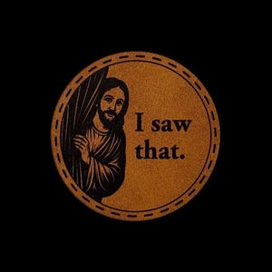 May include: A circular brown leather-like coaster featuring a black engraved illustration of a bearded figure peeking from behind a curtain. The text "I saw that." is printed in black to the right of the figure. The edge has a stitched border.