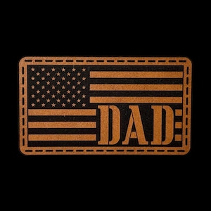 May include: A rectangular leather patch featuring a laser-engraved American flag design. The word DAD is prominently displayed in large capital letters across the stripes on the right side. The patch has a stitched border detail around the edge.
