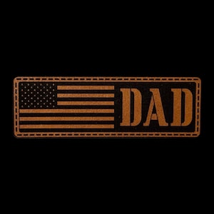 May include: A rectangular brown leather patch featuring a laser-engraved American flag on the left side and the word DAD in bold, capitalized letters on the right side. The patch has a stitched border detail around the entire perimeter.