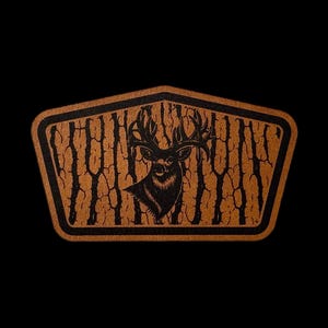 May include: A pentagonal patch featuring a tan, textured background with a black, laser-engraved graphic of a deer head with large antlers. The design includes vertical, tree-like bark patterns surrounding the deer, all enclosed by a thick black border.