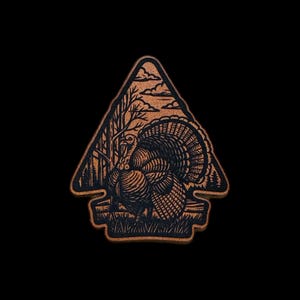 May include: A wooden turkey-shaped emblem with a detailed illustration of a turkey in a forest scene. The emblem is brown with a black outline, and the background is black. The emblem has a triangular shape with a decorative border.