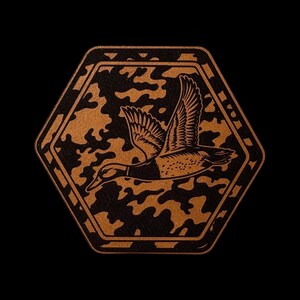 May include: A hexagonal patch featuring a copper-colored flying duck silhouette centered against a black and copper camouflage pattern. The design is enclosed within a thick copper border, creating a sharp contrast against the dark background.