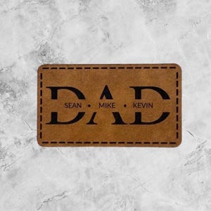 May include: A rectangular brown leather patch with rounded corners and black stitched borders. The word DAD is printed in large black letters, with the names SEAN, MIKE, and KEVIN printed in smaller black text between the letters of DAD.