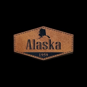 May include: A brown leather patch with the word "Alaska" in bold black letters, the state's outline, and the year "1959". The patch has a stitched border and a black background.