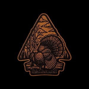 May include: A tan, arrowhead-shaped patch featuring a black engraved illustration of a wild turkey standing in a grassy field with trees and clouds in the background. The design is enclosed within a thick black border, creating a high-contrast graphic.