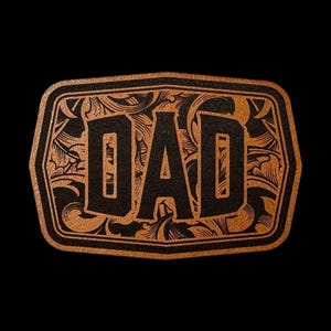 May include: A rectangular belt buckle with rounded corners featuring a copper-colored finish. The center displays the word DAD in bold black lettering, surrounded by an intricate black engraved floral scrollwork pattern against the copper background.