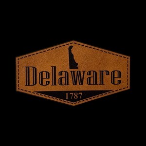 May include: A brown hexagonal leather patch featuring a stitched border. The text Delaware is printed in black, with the silhouette of the state above the letters. The year 1787 is printed in black at the bottom center of the patch.