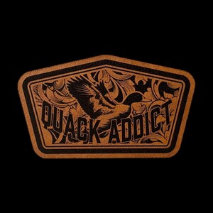 May include: A pentagonal patch featuring a tan leather-like surface with a black engraved design. The graphic depicts a flying duck surrounded by intricate scrollwork patterns and the bold, capitalized text QUACK ADDICT centered below.