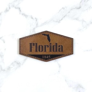 May include: A brown leather patch with the word "Florida" in a bold font, the outline of the state, and the year "1845". The patch has a stitched border and a geometric shape.