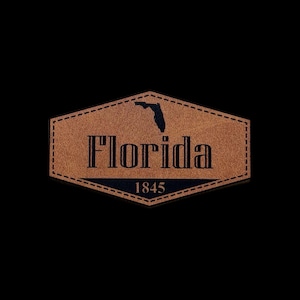 May include: A brown leather patch with the word "Florida" in bold black letters, the silhouette of Florida, and the year "1845". The patch has a stitched border and is set against a black background.