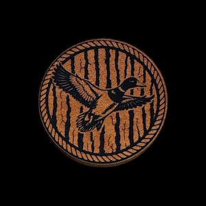 May include: Circular wooden coaster with a detailed laser-etched design. The coaster features a flying duck silhouette in black against a background of vertical lines, all framed by a rope-like border. The wood has a warm, brown tone.