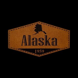 May include: A hexagonal brown leather patch featuring a black silhouette of the state of Alaska, the word Alaska in a serif font, and the year 1959 printed in black. The patch has a decorative stitched border around the entire perimeter.