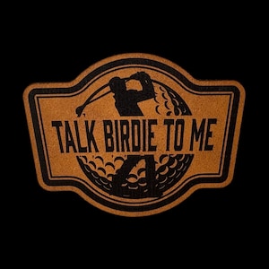 May include: A brown, textured, shield-shaped patch featuring a black graphic of a golf ball and a golfer mid-swing. The text "TALK BIRDIE TO ME" is printed in bold, black, sans-serif capital letters across the center of the design.