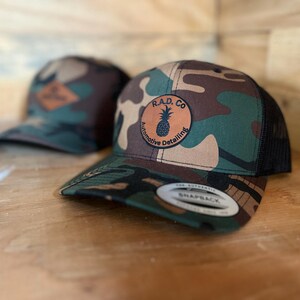 Op de afbeelding: Een camouflage trucker-pet met een zwart mesh-achterpaneel. De pet heeft een bruin leren patch met de tekst "R.A.D. CO. Automotive Detailing" en een ananas-afbeelding.