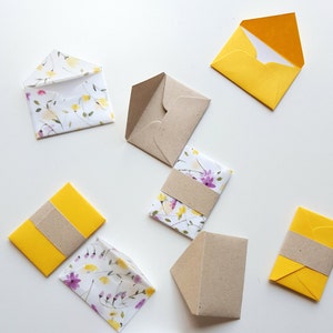 May include: Assortment of small envelopes in various colors and patterns. Some envelopes are yellow, some are brown, and some have a floral pattern. The envelopes are all closed and are arranged on a white surface.