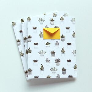 May include: A stack of three white notebooks with a repeating pattern of potted plants. The notebooks have a yellow envelope on top.