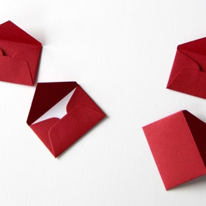 May include: Five red envelopes of various sizes are arranged on a white surface. The envelopes are all closed and have a white interior.