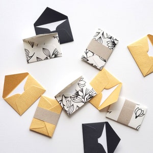 May include: A collection of small envelopes in various colors and patterns, including black, white, and gold. The envelopes are decorated with floral designs and are perfect for sending notes or small gifts.