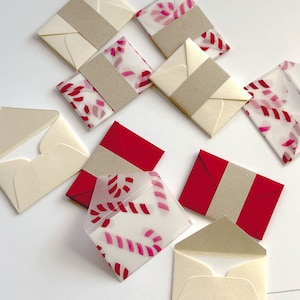 May include: An assortment of small, cream-colored paper envelopes, some with red and pink candy cane patterned inserts. Several envelopes are sealed with a tan paper band, while others are open. The background is white.