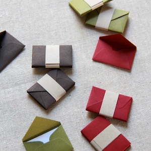 May include: A collection of small, folded envelopes in various colors, including green, red, and brown. Each envelope has a brown paper flap with a white paper strip for sealing.
