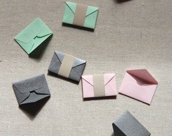 May include: Assortment of small envelopes in various colors, including pink, green, and gray. The envelopes are folded and have a flap on the top. They are arranged on a textured beige surface.