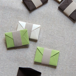 May include: Six small envelopes with different colored paper and brown paper flaps. The envelopes are on a light brown linen fabric.