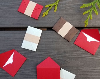 May include: Six small envelopes in various colors, including red, brown, and white, with a brown paper insert inside each envelope. The envelopes are arranged on a dark gray wooden surface.