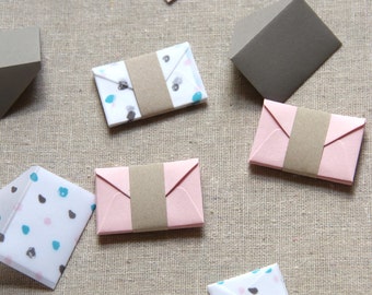 May include: A collection of seven small envelopes with different patterns and colors. The envelopes are made of paper and have a brown paper flap. The envelopes are arranged on a light brown fabric background.