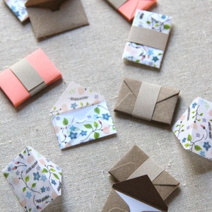 May include: A collection of eight small envelopes in various patterns and colors. The envelopes are made of paper and have a brown paper flap. Some of the envelopes have a floral pattern, while others are solid pink or brown.